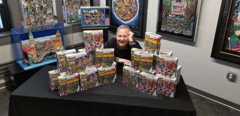 Charles Fazzino sitting at a table covered with boxes of puzzles from his new puzzle line.