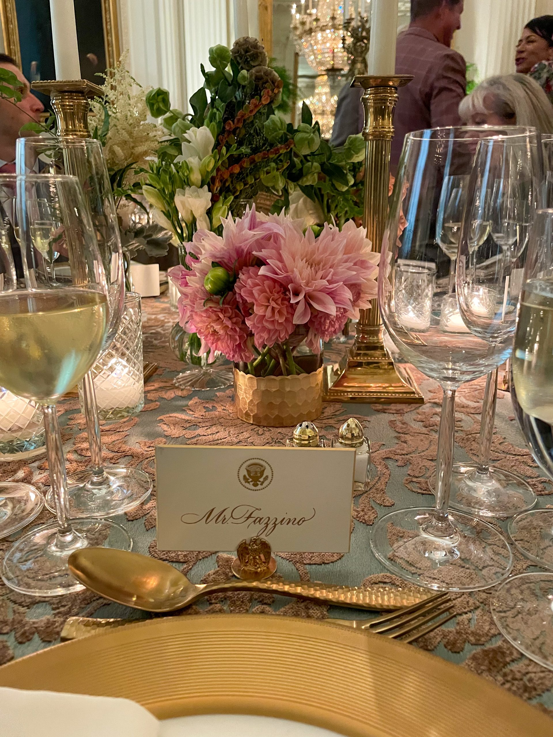 La tarjeta de lugar que dice Sr. Fazzino sentado frente a un plato con borde dorado, una cuchara dorada, copas de vino y hermosas flores rosas.