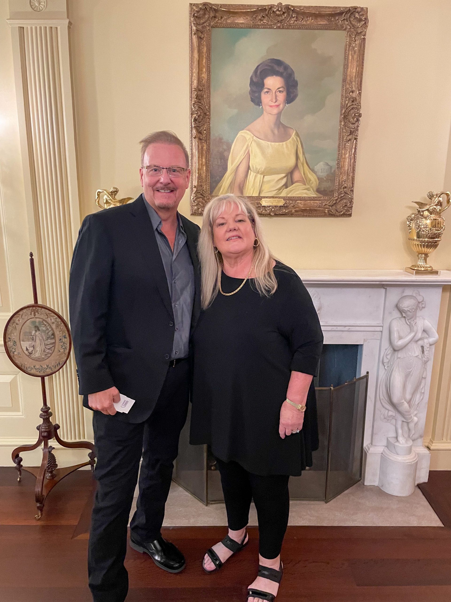 Charles and his wife stand in front of a fireplace and painting of a first lady in the white house