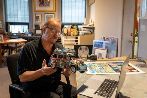 Fazzino sitting in his studio, holds up his VW bug car sculpture that is adored with 3D elements